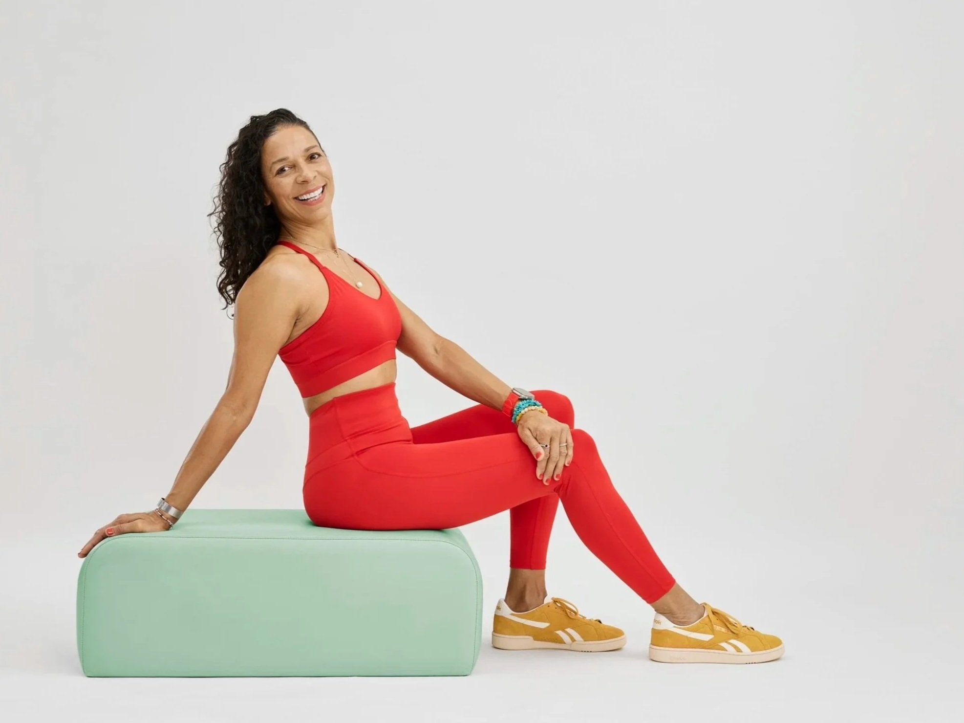 LaBriece wearing a red sports bra, red leggings, and yellow sneakers, sitting on a light green reformer bench against a plain white background, smiling at the camera.