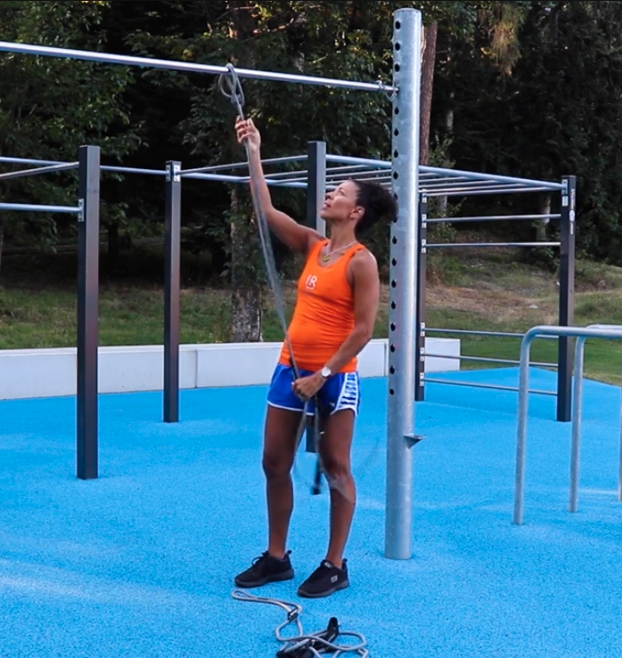A woman standing on a blue outdoor gym floor, holding a climbing rope, preparing to climb a horizontal bar in a fitness park with green trees in the background.