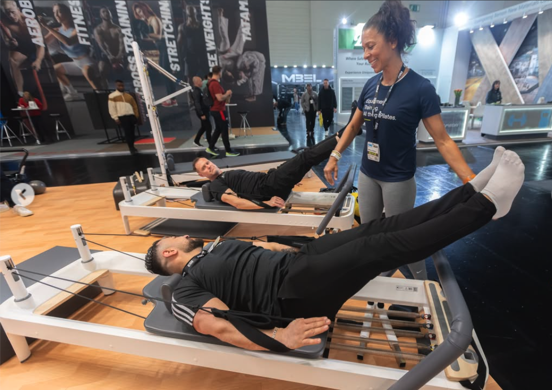 A woman assisting two men exercising on specialized Pilates reformer machines at a fitness expo.