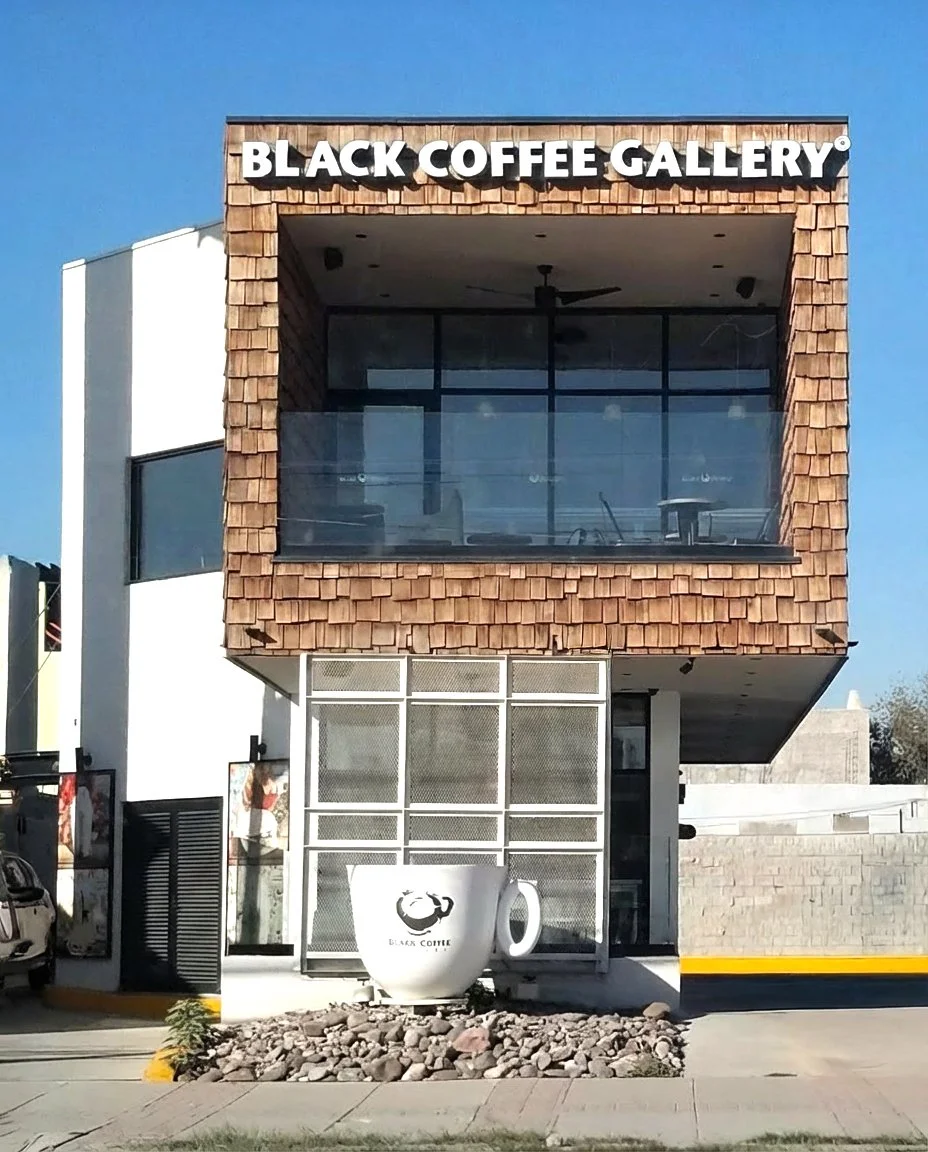 A two-story building with a sign that reads 'Black Coffee Gallery' on the top. The building has a large mug-shaped decoration outside with 'Black Coffee' and a coffee cup logo on it. The building features modern architecture with wood shingles on the upper level and glass windows.