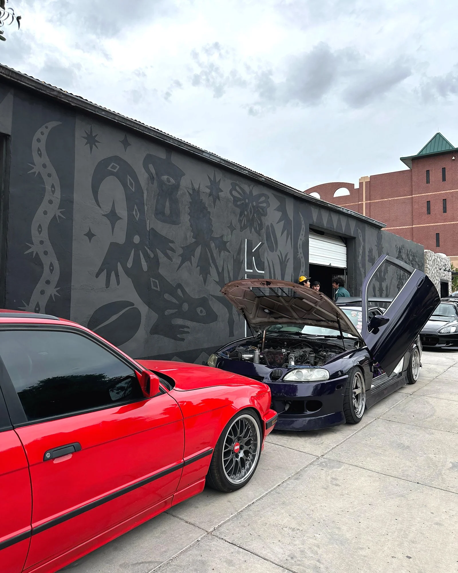 A red sports car parked next to a purple sports car with its hood open, in front of a mural and a brick building.