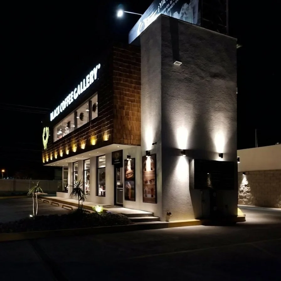 Night view of a building with illuminated signs, including a gallery and cafe, with outdoor lighting, plants, and a parking lot.