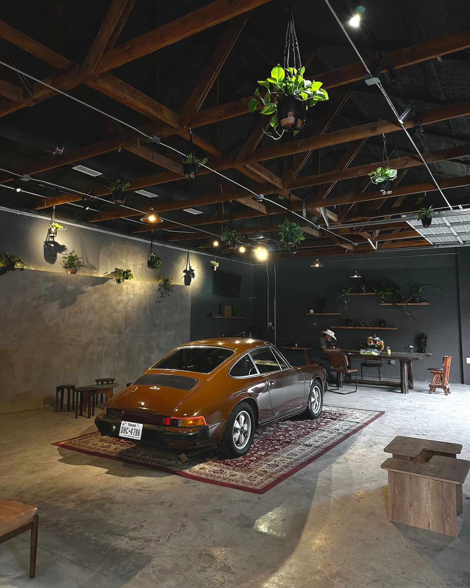 Vintage car parked inside a stylish industrial-style room with hanging potted plants, wooden elements, and a red patterned rug.