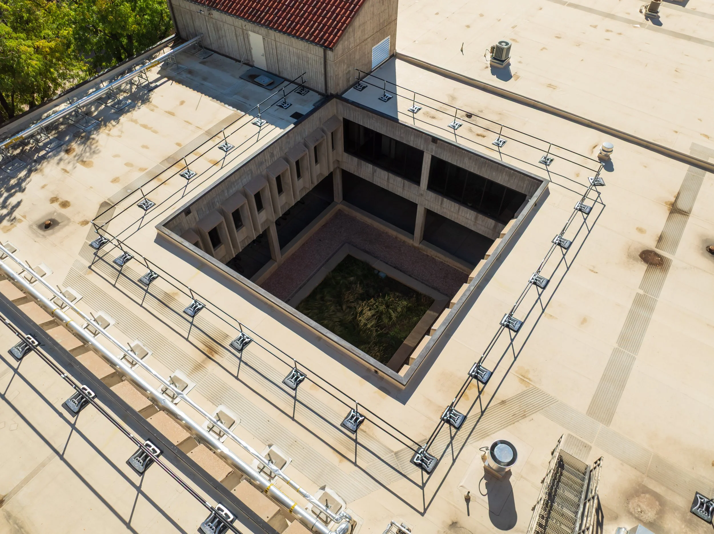 Aerial view of a rooftop with an open courtyard in the center, surrounded by a railing and ventilation units, with some trees visible at the edge of the roof.