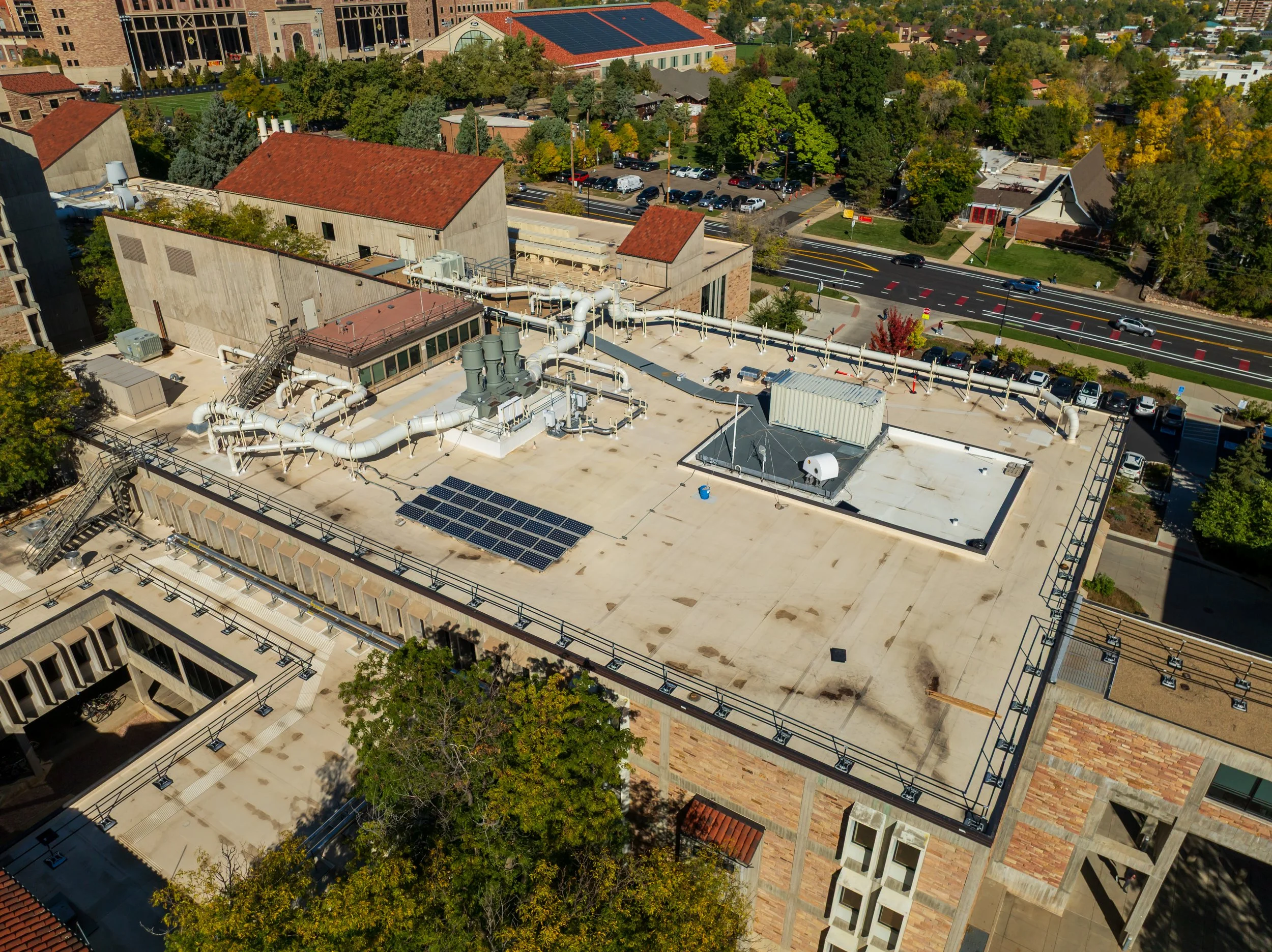 Aerial view of a building rooftop with HVAC equipment, pipes, solar panels, and a small rooftop structure, with streets and trees in the background.