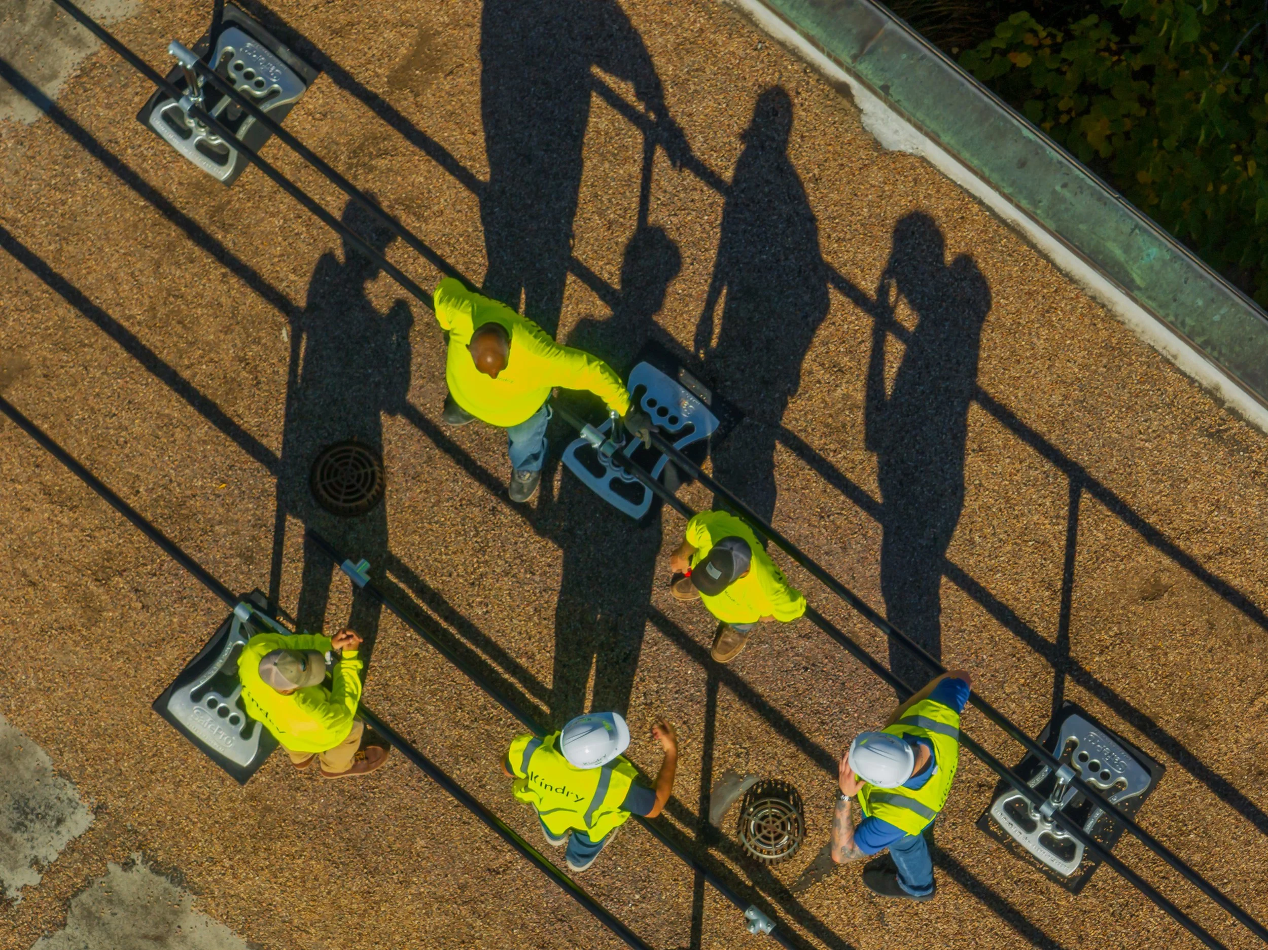 Aerial view of five construction workers wearing yellow safety vests and helmets, working on electrical or utility equipment on a rooftop, with shadows cast on the gravel surface.