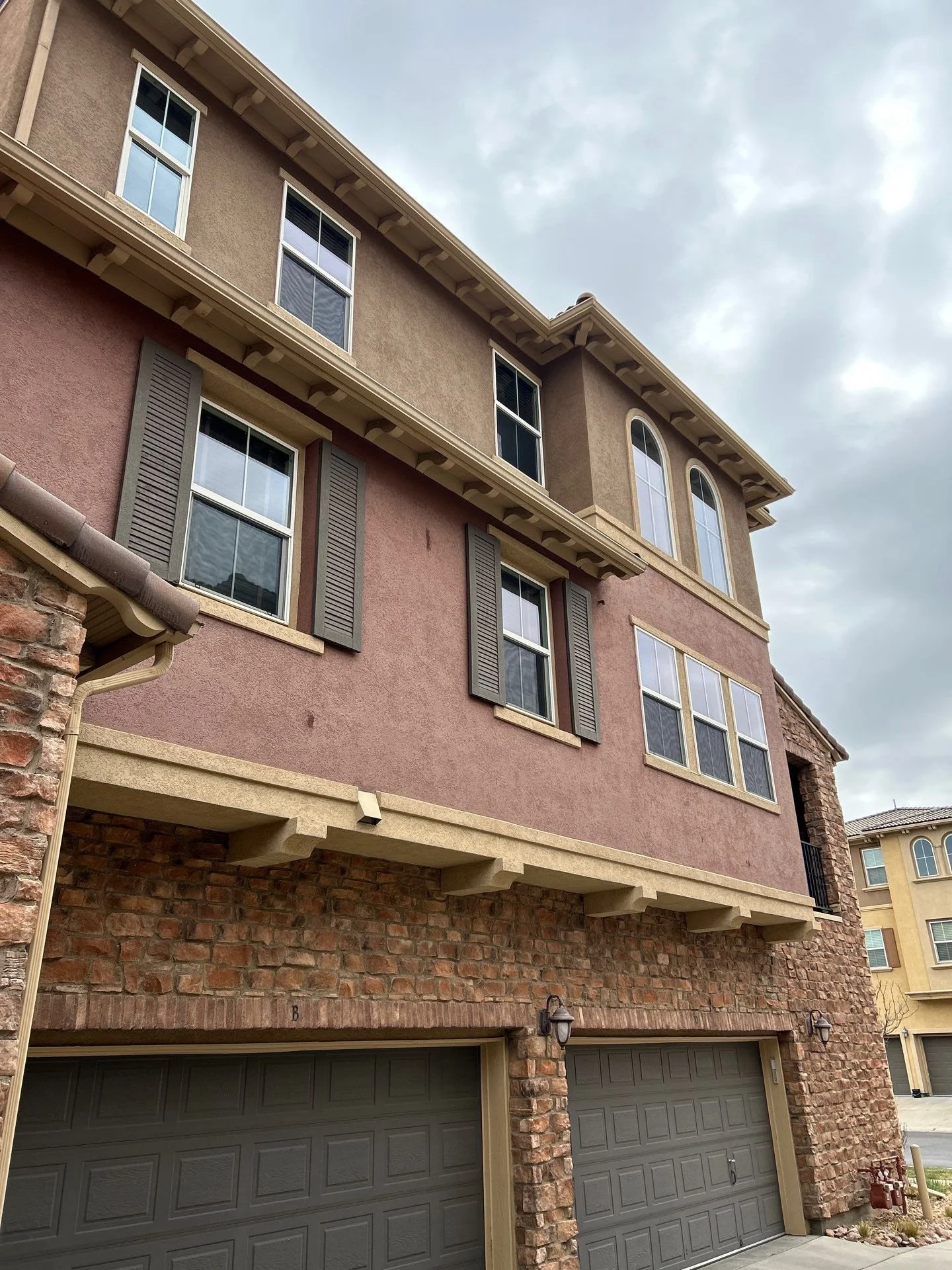 Four-story residential building with stone and stucco exterior, attached two-car garage, multiple windows with shutters, and decorative architectural elements. Overcast sky.