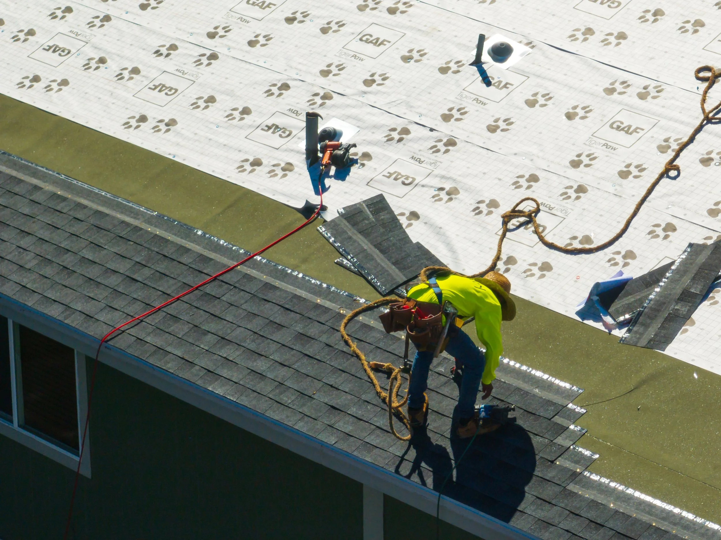 Worker installing or repairing roof shingles on a house, with roofing materials and tools around him.