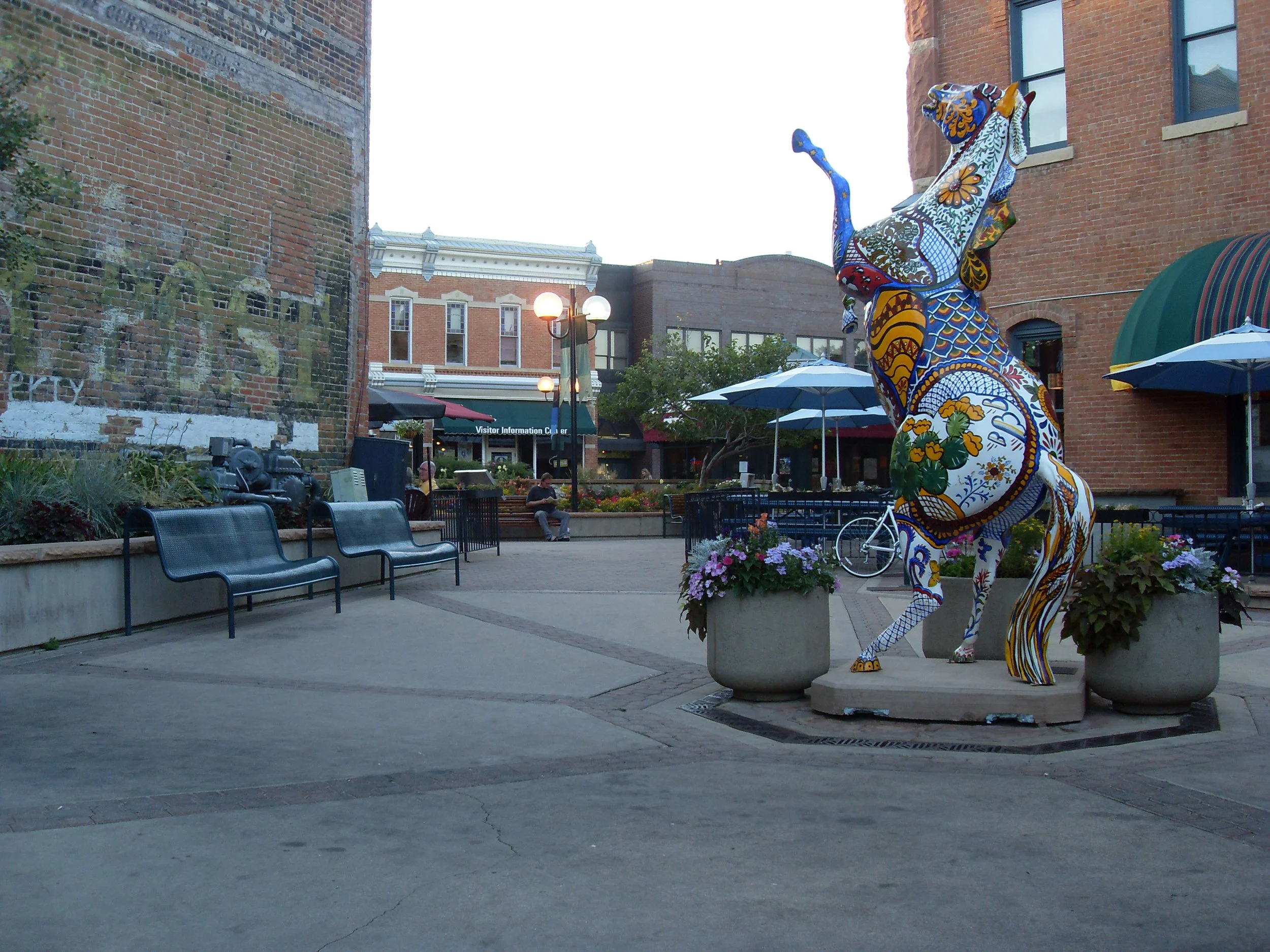Colorful ceramic sculpture of a giraffe in an outdoor urban plaza with brick buildings, benches, and a few people sitting, along with flower planters and umbrellas.