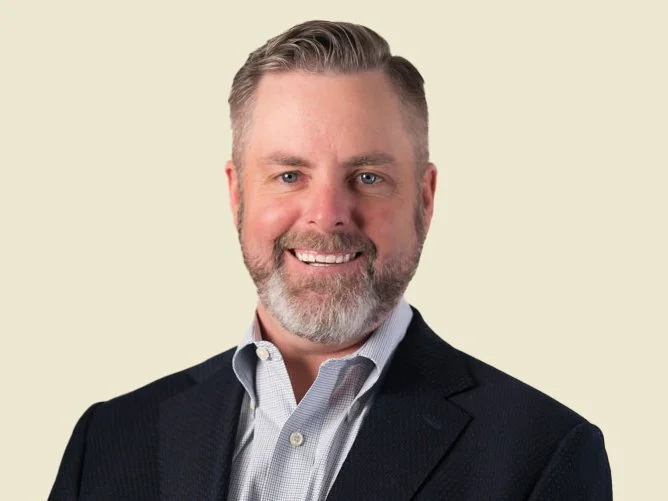 Headshot of a man with short gray hair, beard, wearing a dark suit jacket and a light-colored collared shirt, smiling against a plain light background.