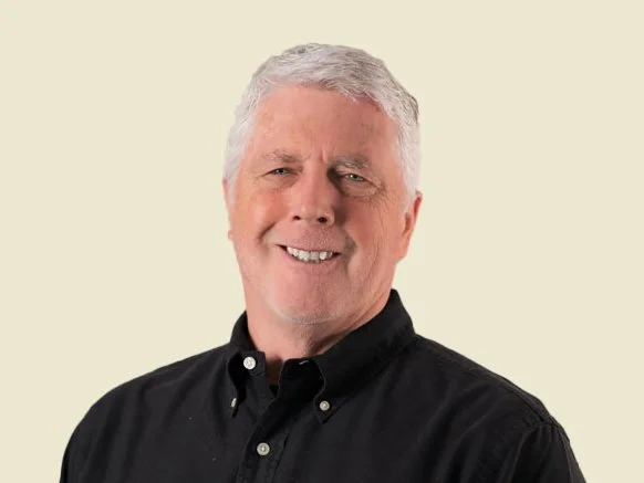 A smiling older man with gray hair and a beard, wearing a black button-up shirt, posed against a light beige background.