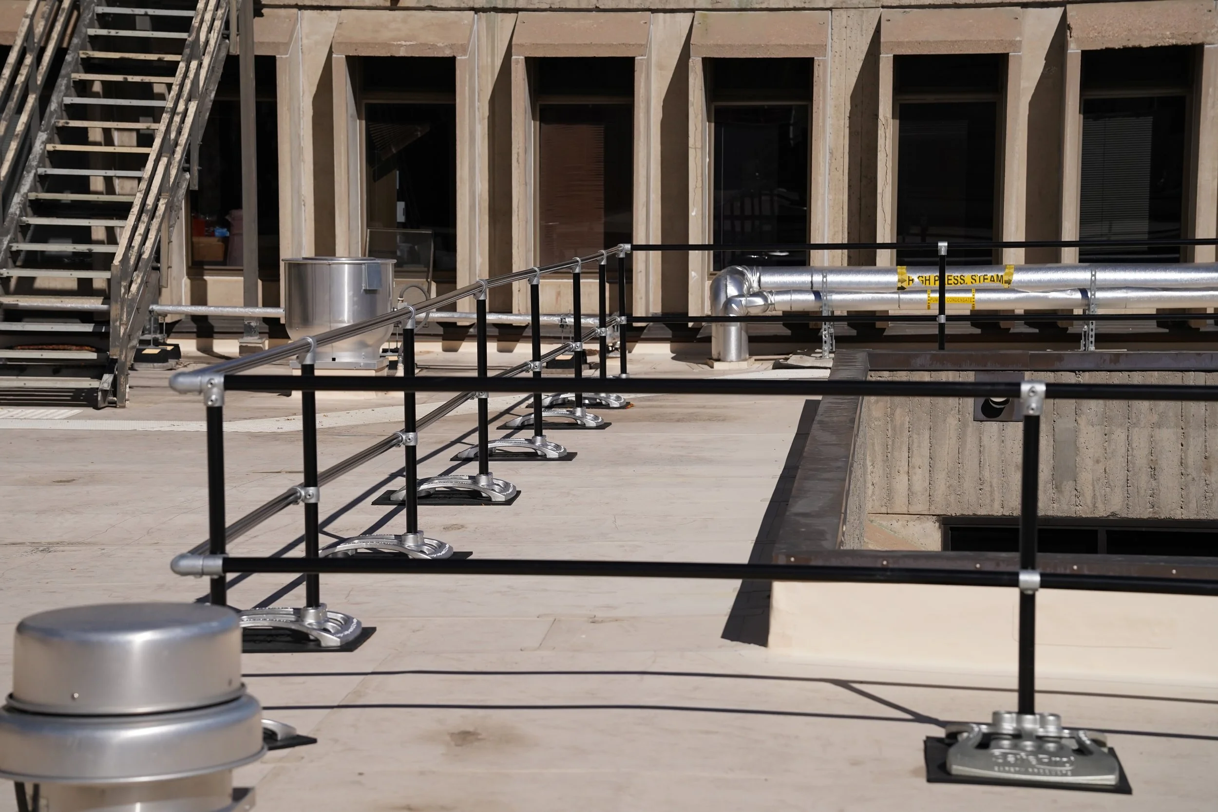 Empty rooftop area with construction barriers, metal piping, and stairs in front of a building with large windows.