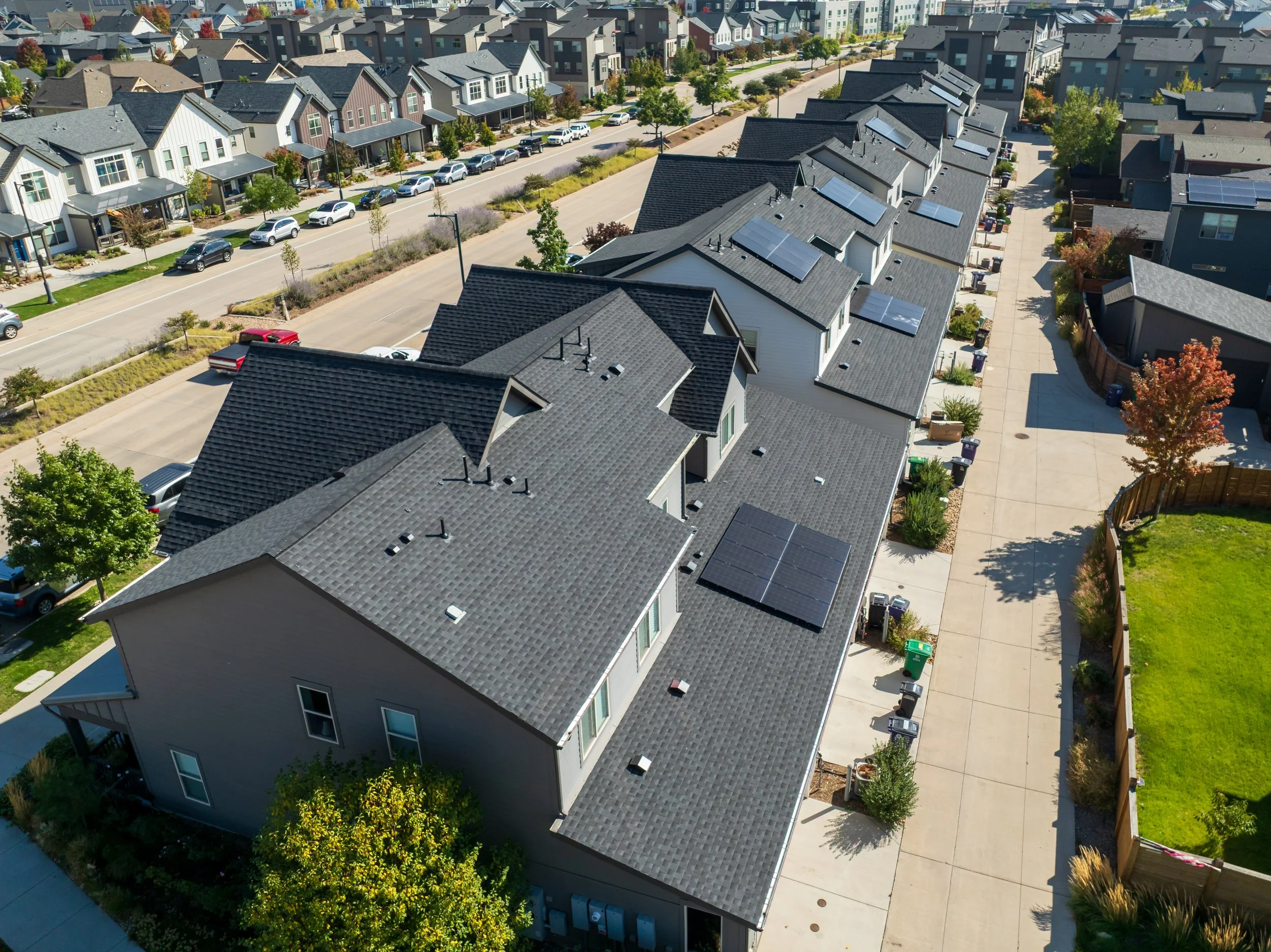 Aerial view of a row of modern houses with solar panels on their rooftops, parked cars along the street, and a sidewalk with trees and shrubs in a residential neighborhood.