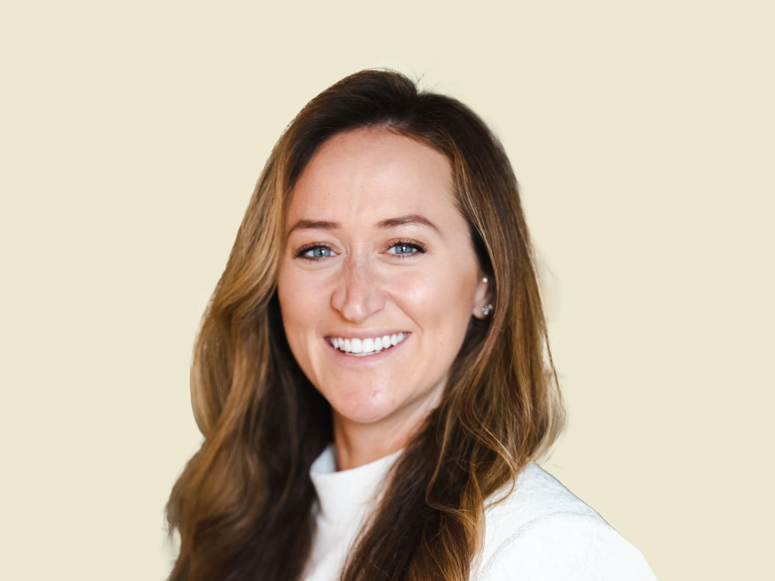 A smiling woman with long brown hair, blue eyes, and earrings wearing a white top, posing against a plain light-colored background.