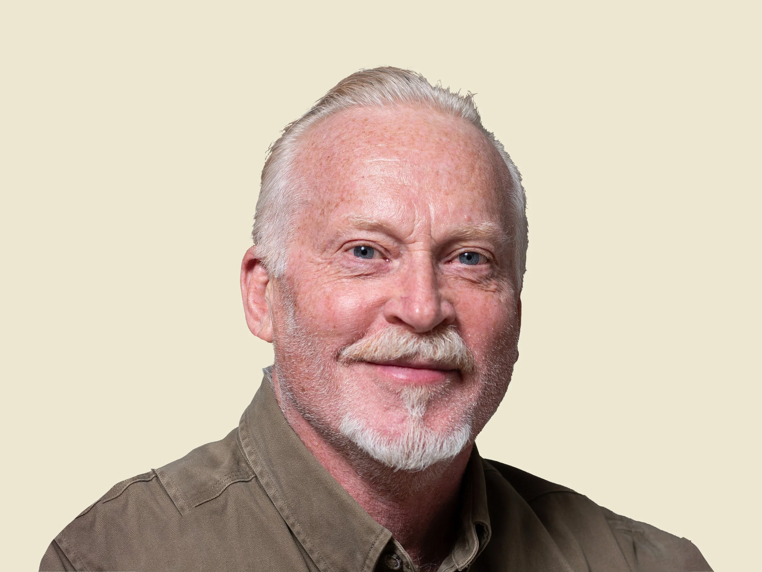 Close-up portrait of an older man with white hair, a beard, and mustache, wearing a brown shirt, smiling slightly, against a plain light background.
