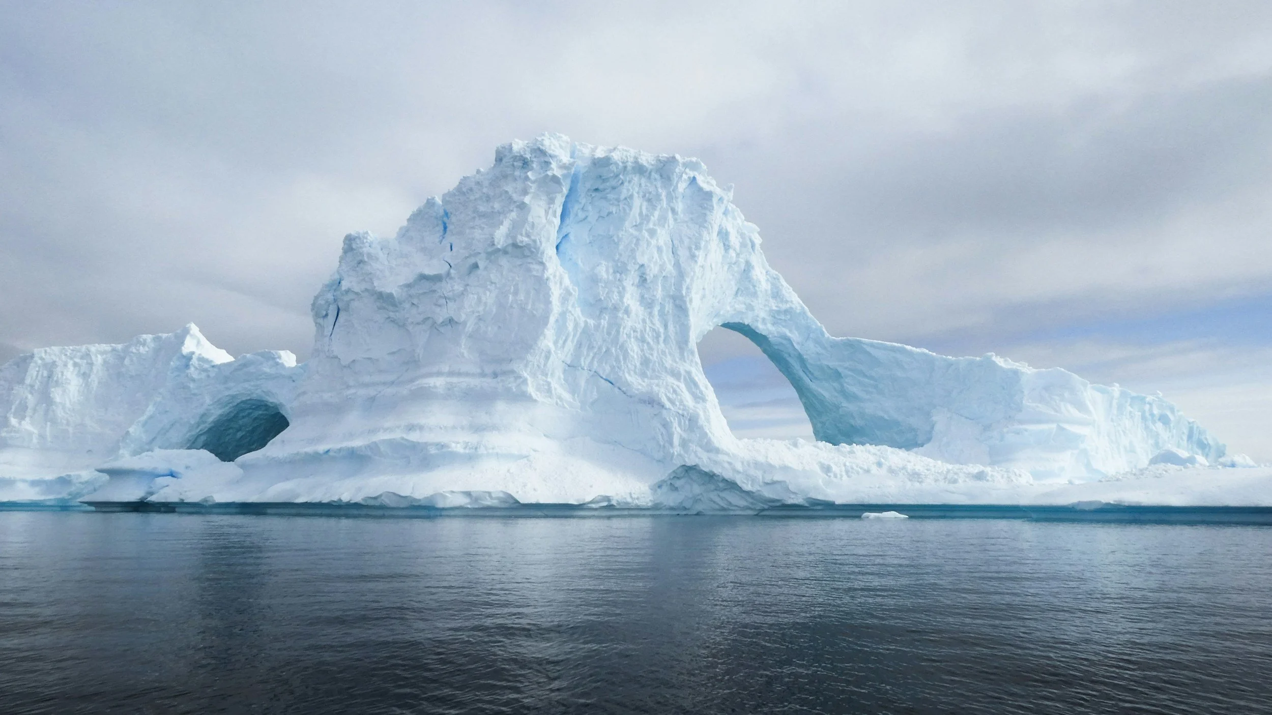 Large iceberg with natural arch formation floating in the ocean under a cloudy sky.