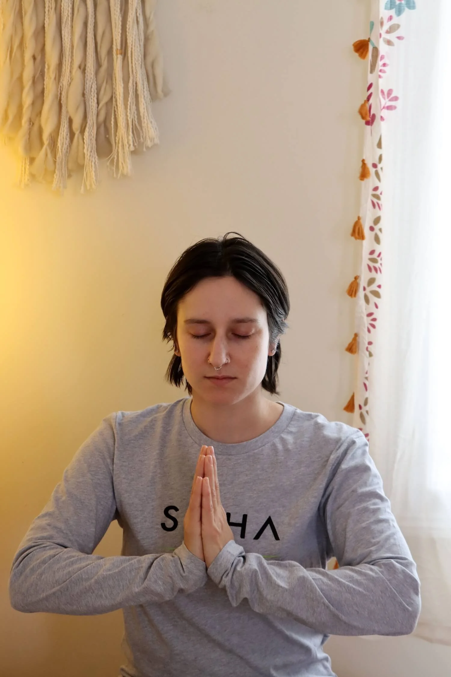 A woman with short dark hair wearing a Saha Yoga long sleeve shirt sits with her hands together