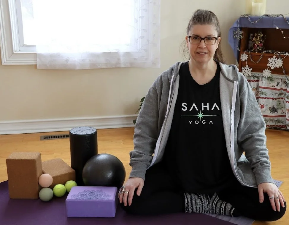 A woman wearing a Saha Yoga shirt sits on the floor surrounded by yoga mats and clocks