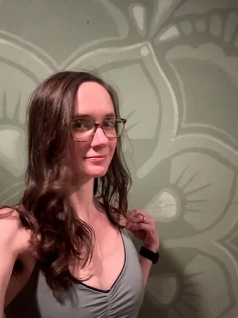 A Woman with long brown hair and glasses stands in front of a wall with a flower painted on it