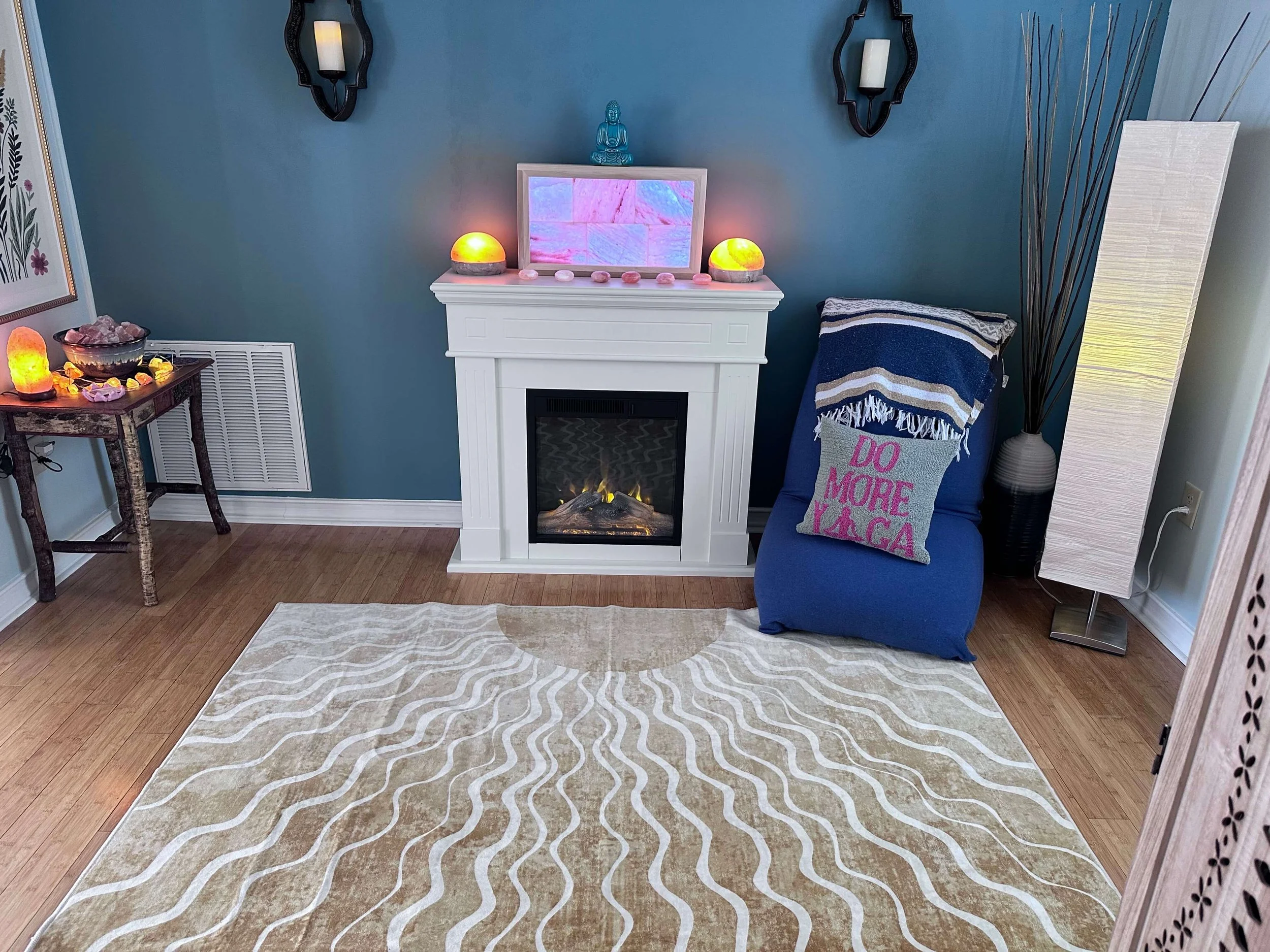 Yoga studio room with light colored rug, a white fireplace with salt lamps, a blue chair and a pillow that says "do more yoga"