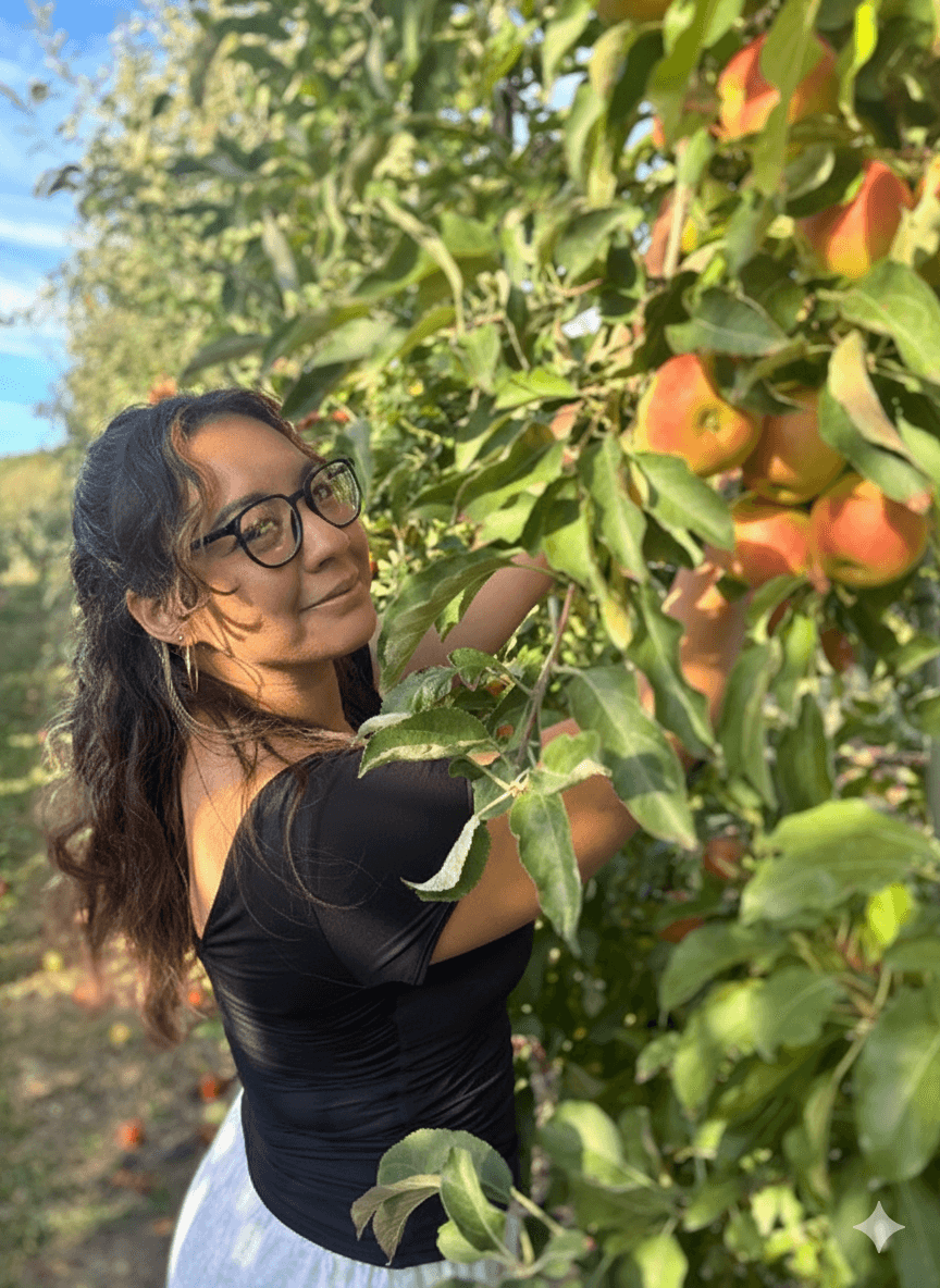 A dark haired woman with glasses stands near an apple tree