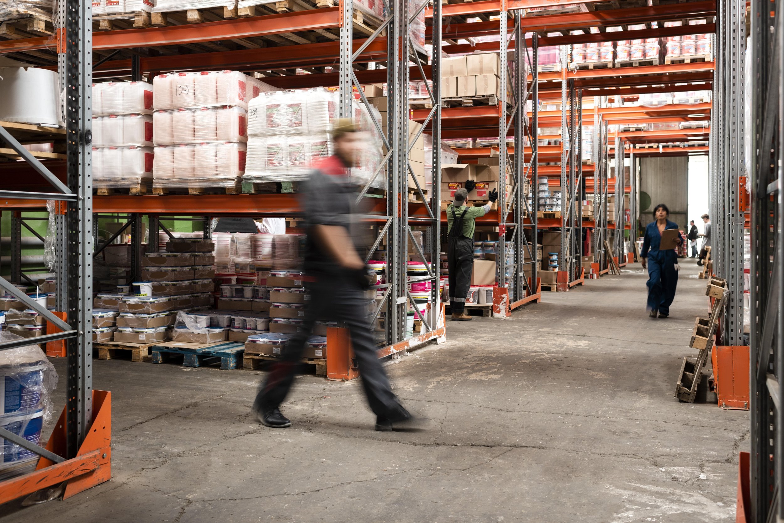 Inside a warehouse with tall metal shelves stocked with boxes and supplies, and several workers moving or inspecting items.