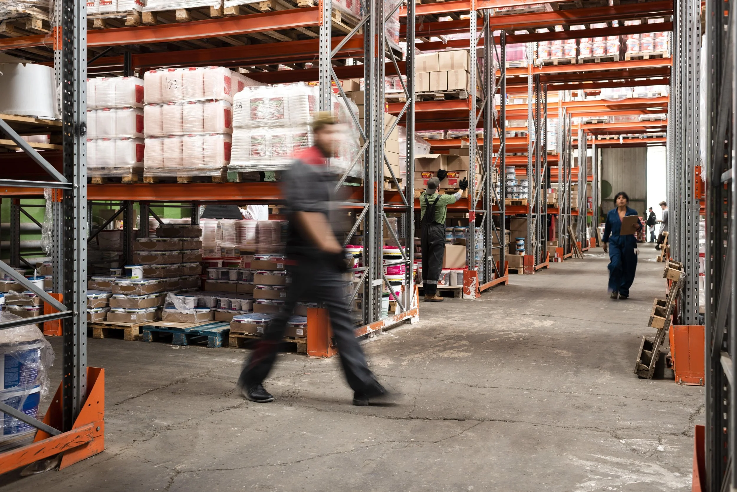 Warehouse with shelves filled with goods, workers organizing supplies, some blurred motion, industrial setting with concrete floor, wooden pallets, and cardboard boxes.