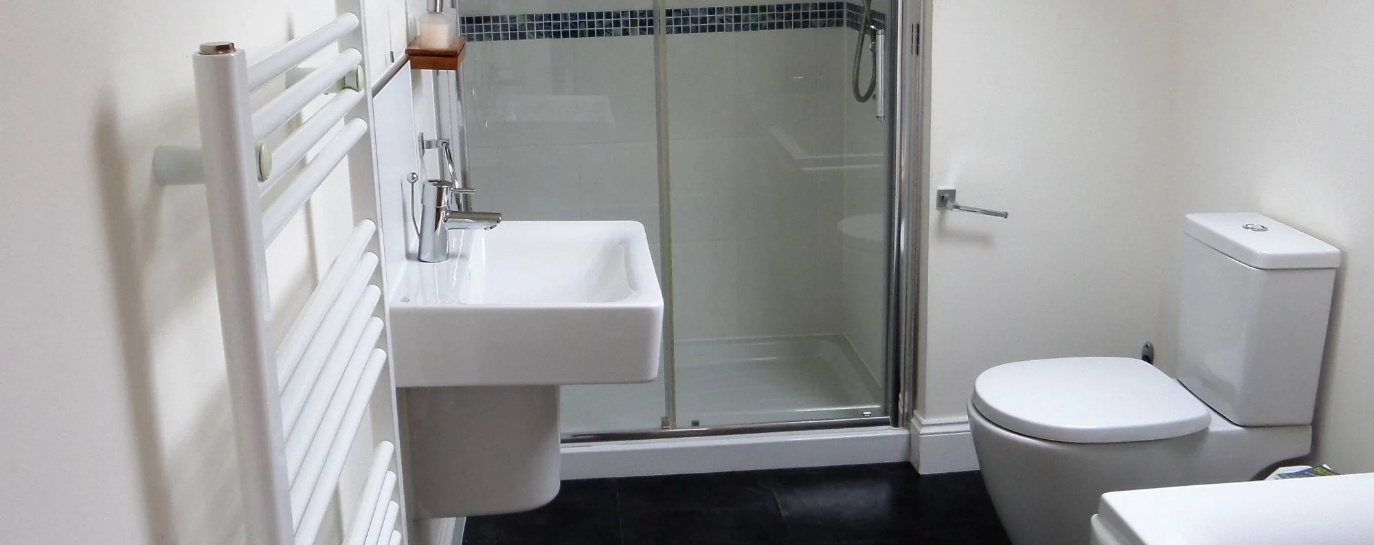 A small bathroom with a white wall-mounted sink with a chrome faucet, a glass shower enclosure, a white toilet, and a towel radiator on the wall.