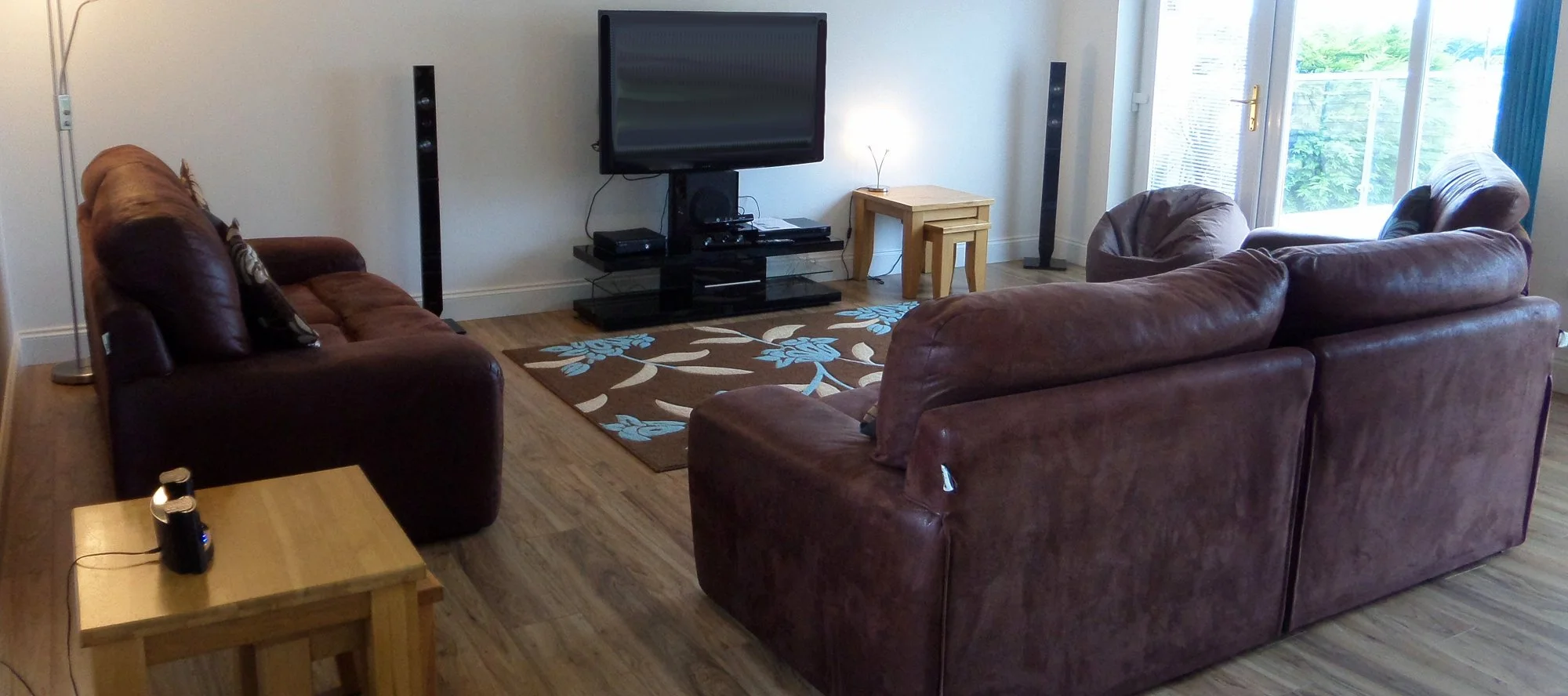 Living room with a brown leather sofa set facing a TV on a black stand, a floral area rug, a small wooden side table, and a large window with blue curtains.