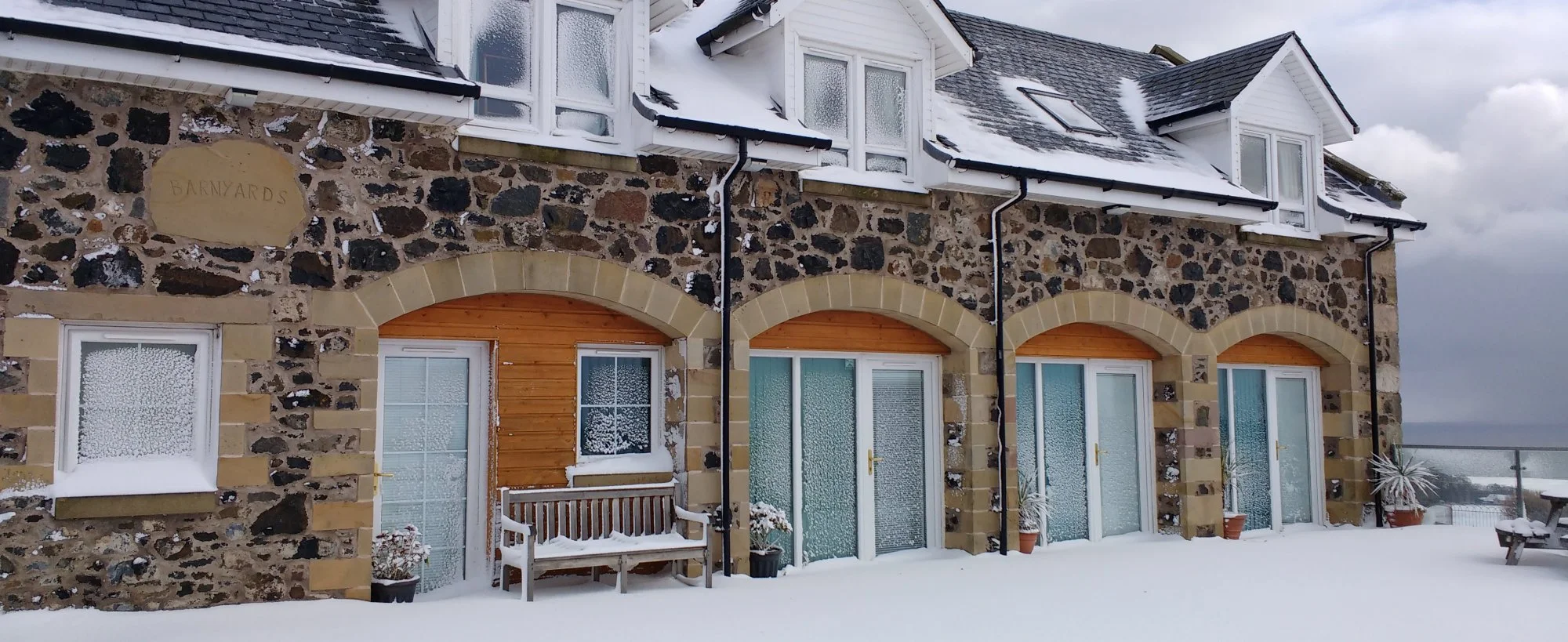 A stone and brick house covered in snow with multiple windows and doors, some with wooden awnings, potted plants, a bench, and a snow-covered outdoor table, under a cloudy sky.