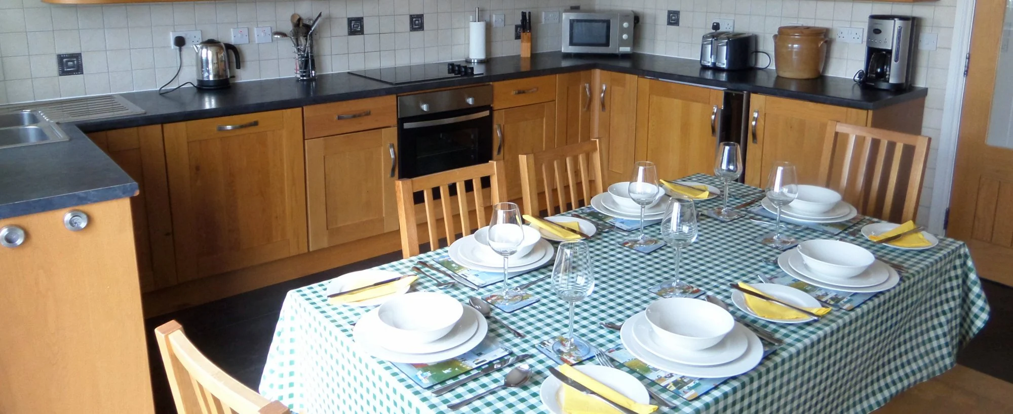 Kitchen with a dining table set for six, featuring white plates, bowls, yellow napkins, and wine glasses on a checkered tablecloth. Kitchen cabinets and appliances including a microwave, toaster, kettle, and coffee maker are visible along the counter