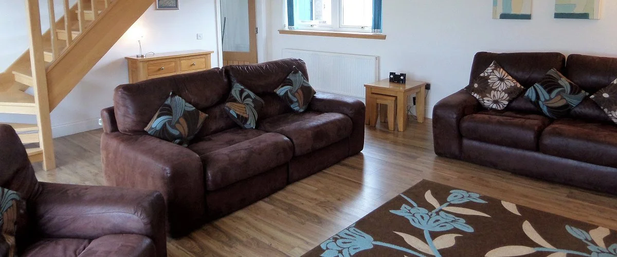 Living room with three brown leather sofas with patterned cushions, wooden flooring, a small wooden side table, a window with blue curtains, a staircase, and a brown floral rug.