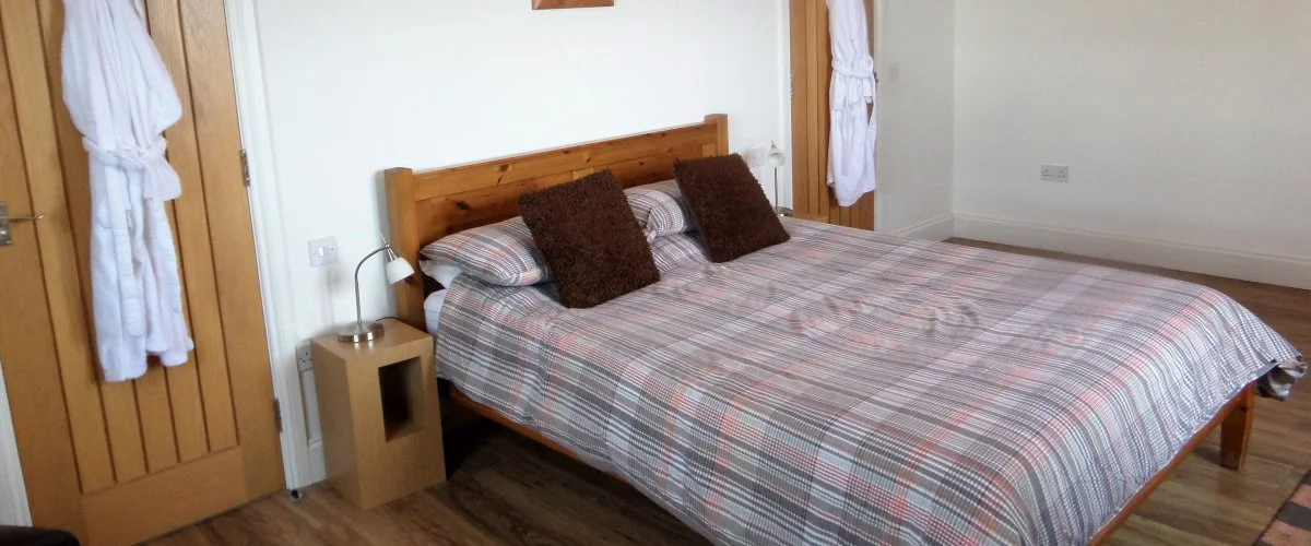 A bedroom with a wooden bed frame, a plaid bedspread, and two brown pillows. There's a nightstand with a small lamp, and white robes hanging on the door and window.
