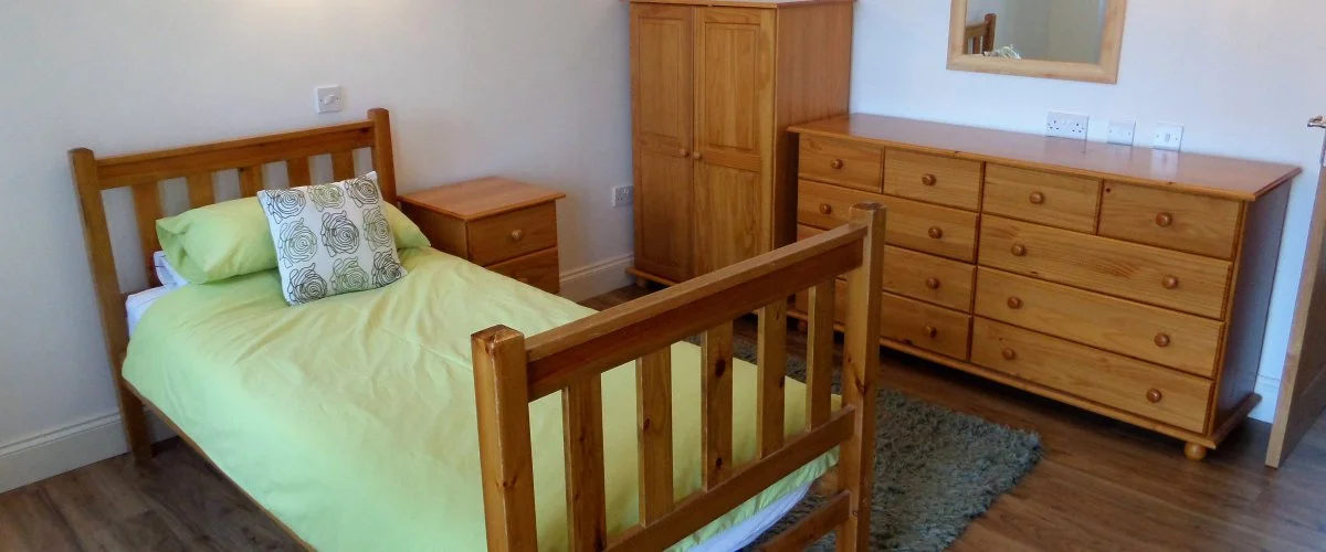 A bedroom with a wooden bed frame, green bedding, two pillows, a wooden nightstand, a wardrobe, a dresser with a mirror, and a small rug on hardwood floor.