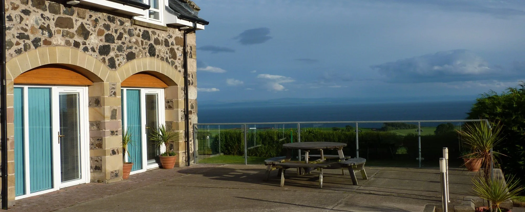 View of a terrace with a wooden bench and potted plants, overlooking a landscape with green fields, trees, and a body of water, under a partly cloudy sky.