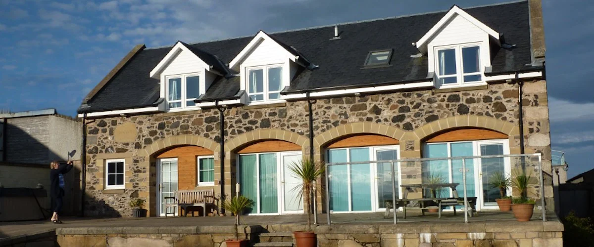 Back view of a stone house with white windows, black roof, and a glass porch, featuring outdoor furniture and potted plants.