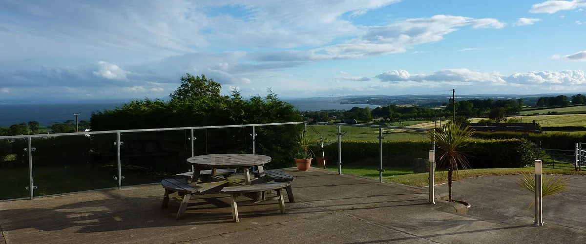 Outdoor patio with a wooden picnic table and bench, potted plants, glass railing, and a scenic view of fields, trees, water, and cloudy sky in the distance.