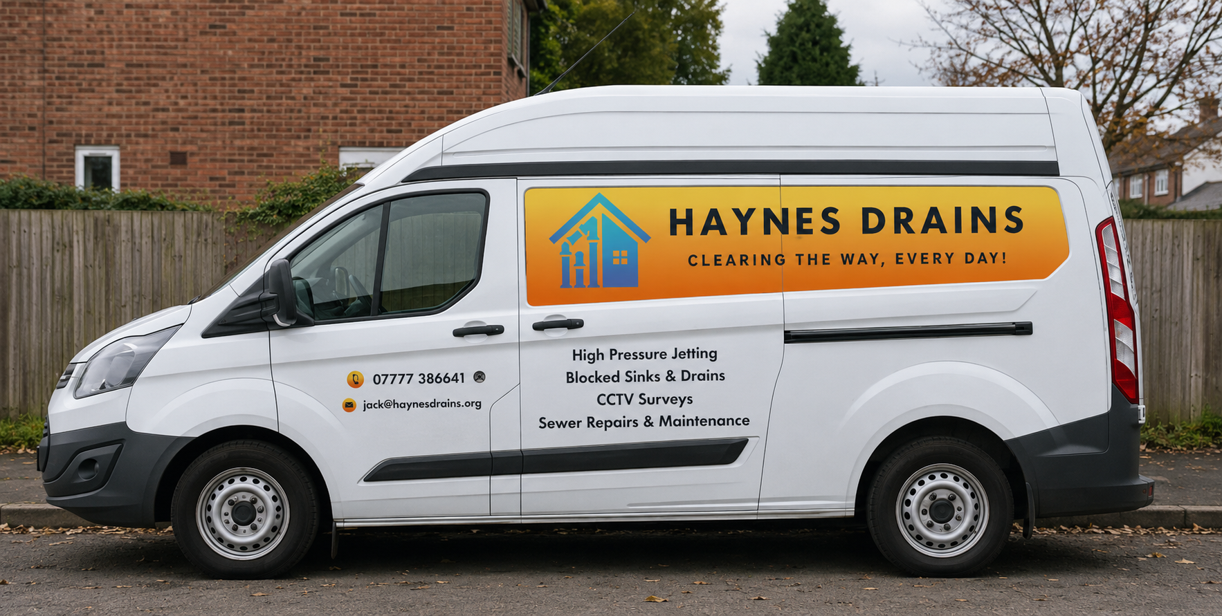 A white service van with Haynes Drains logo and contact information parked on a suburban street with autumn trees and wooden fence in the background.