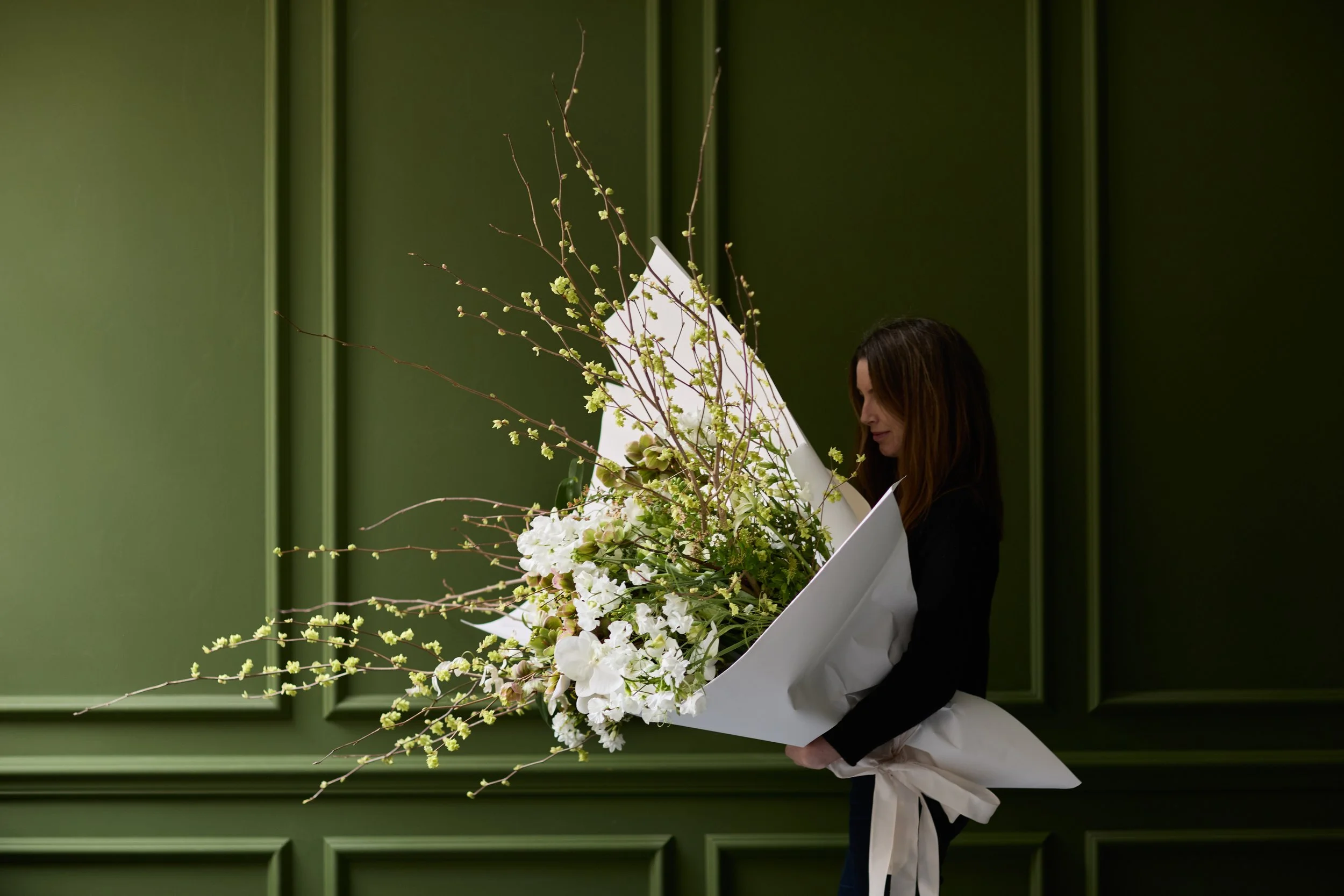 A woman in black clothing holding a large bouquet of white and green flowers against a green paneled wall.
