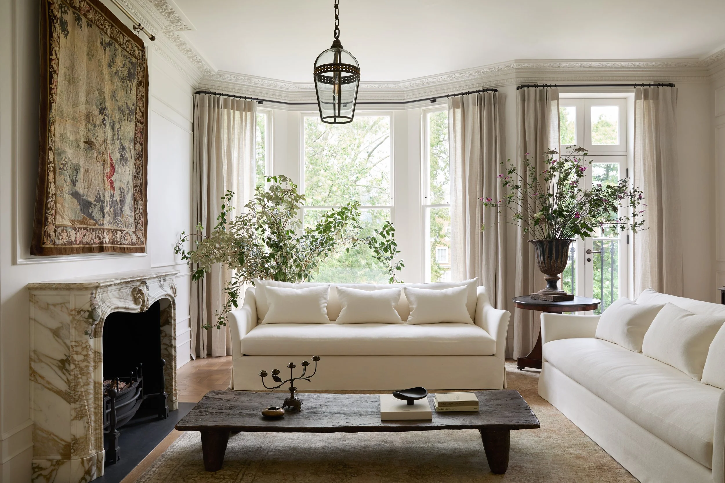 A bright living room with large bay windows, white sofas, a dark wooden coffee table, a decorative vase with flowers, and a tapestry hanging on the wall above a marble fireplace.