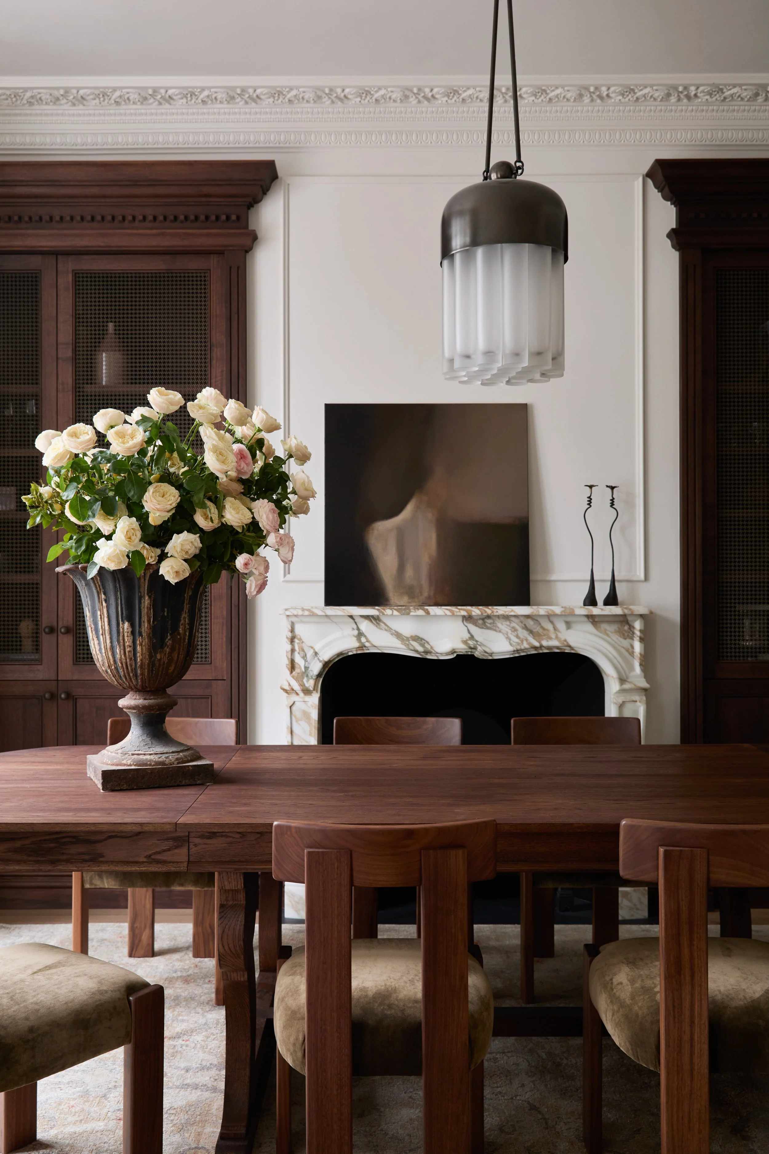 A dining room with a large wooden table, chairs, a vase of white and pink roses, a marble fireplace, artwork, and built-in wooden cabinets.