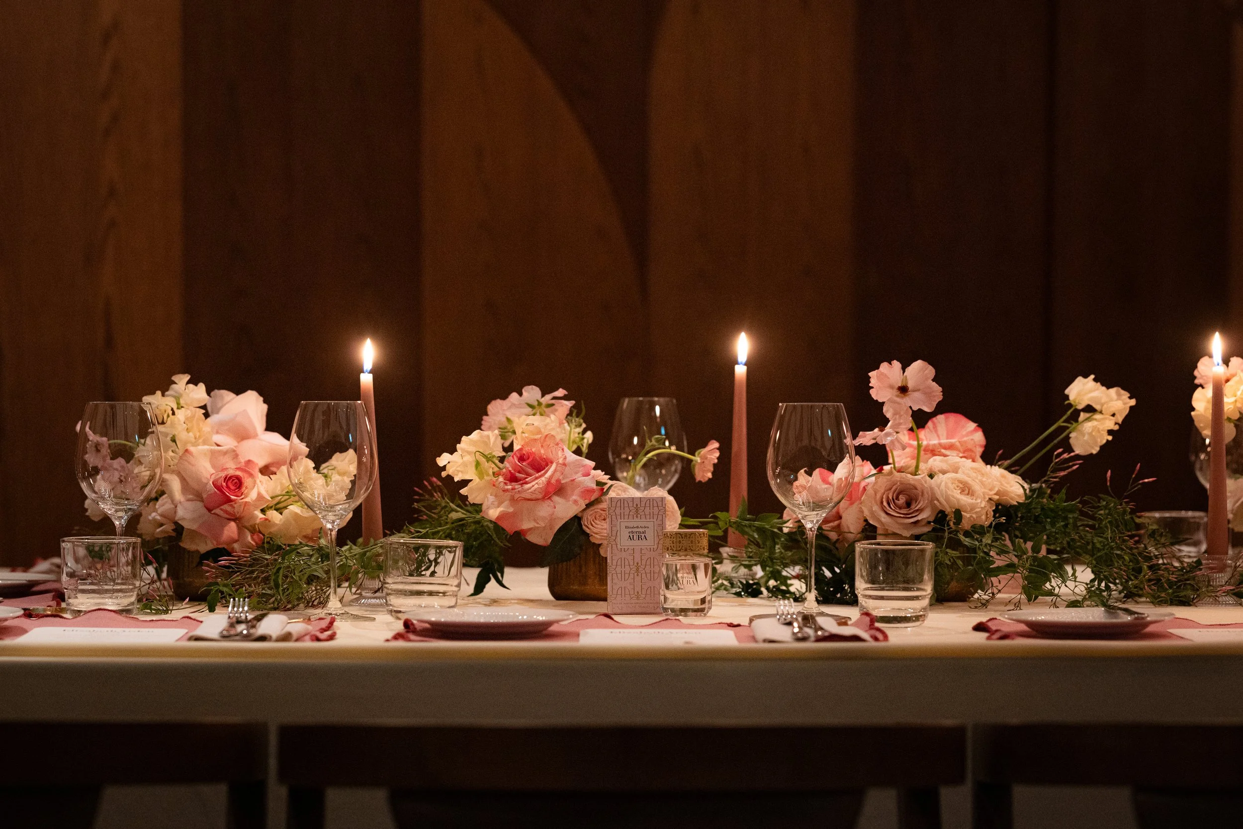 A decorated table for a celebration, featuring a floral centerpiece with pink and white flowers, tall pink candles, and wine glasses. The background is dark wood paneling.