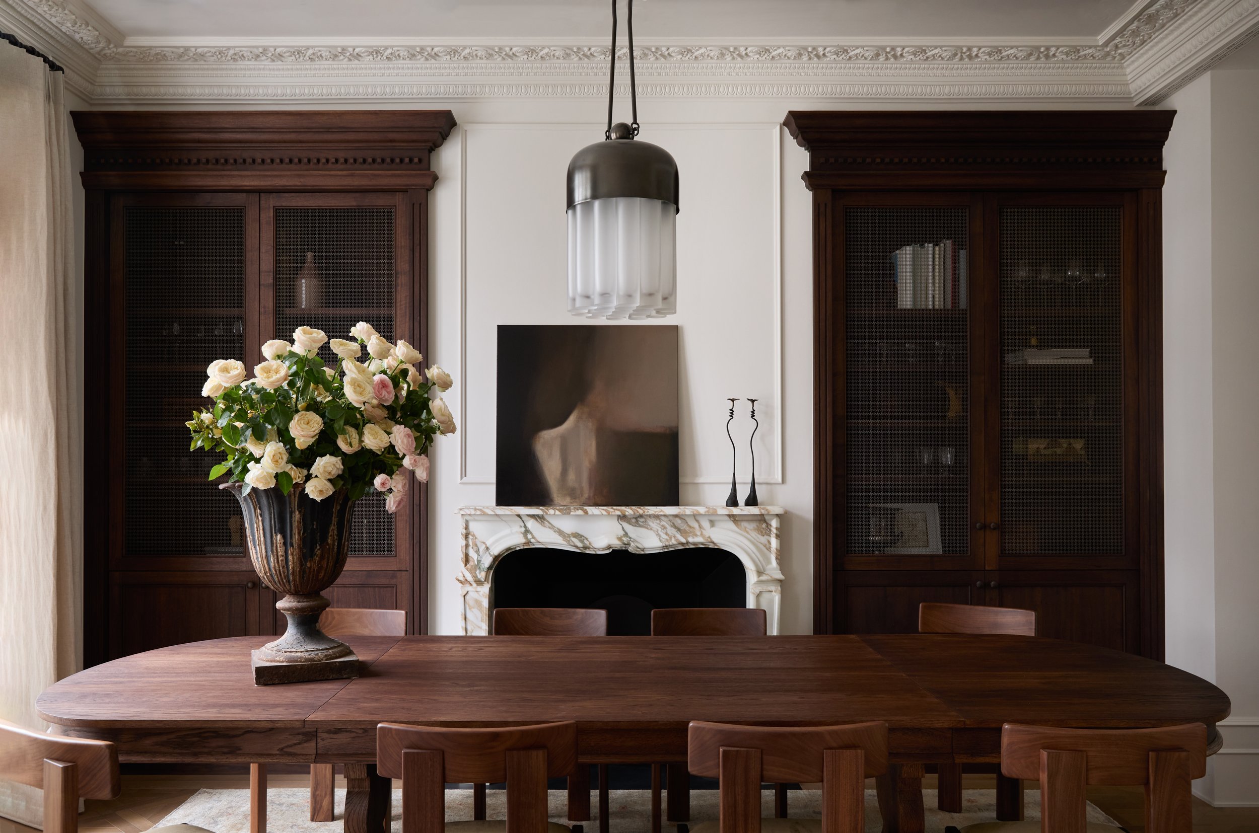 A dining room with a wooden table and chairs, a large vase with pink and white roses, a marble fireplace, dark wooden bookshelves, a TV on the mantel, and a modern pendant light hanging from the ceiling.