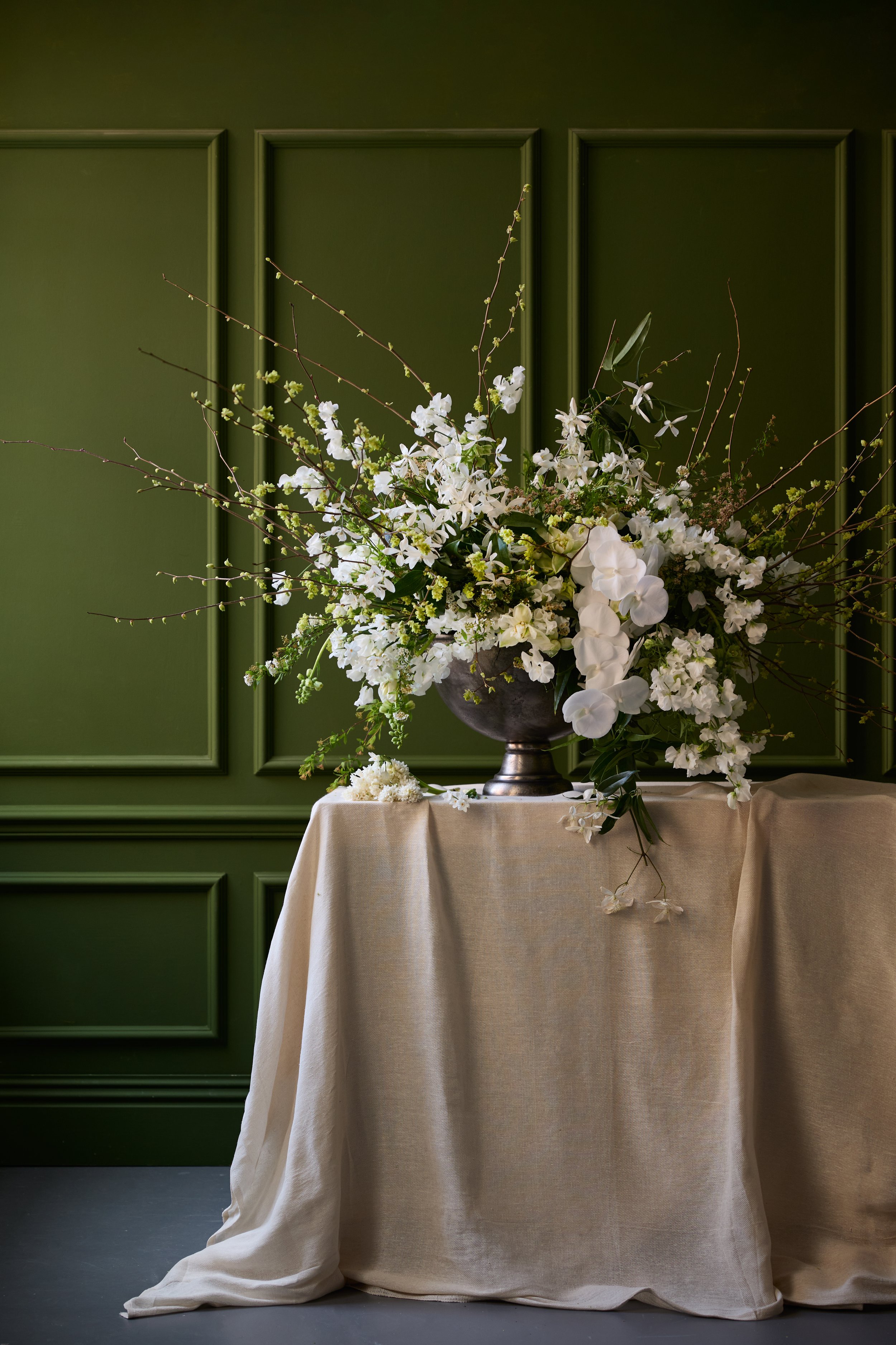 A flower arrangement with white orchids, small white flowers, and green leaves in a large metallic vase on a table with beige cloth, against a dark green paneled wall.