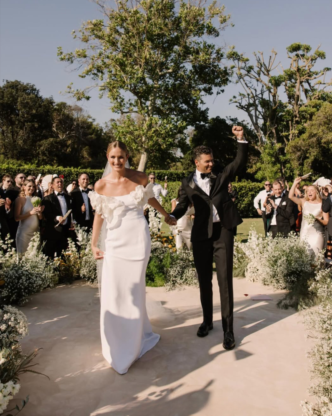 Bride and groom celebrating their wedding outdoors, holding hands, with guests in formal attire clapping and cheering around them.