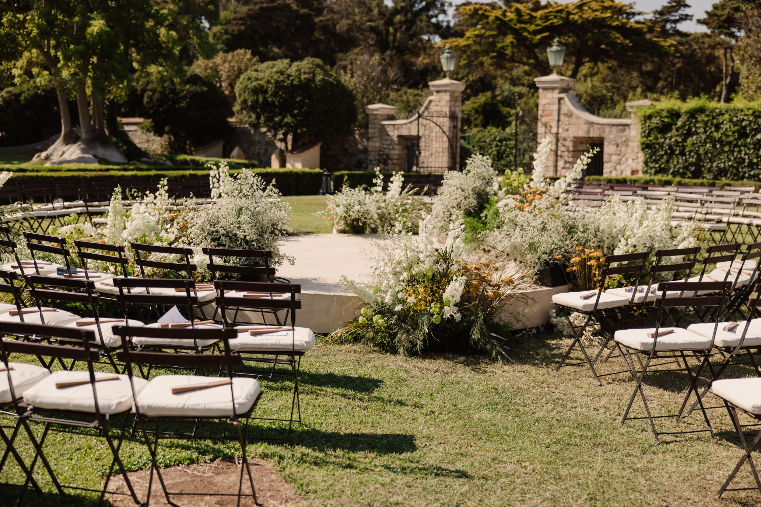 Outdoor wedding or event setup in a garden with chairs arranged around a floral centerpiece on a white platform, surrounded by lush green trees and stone walls.