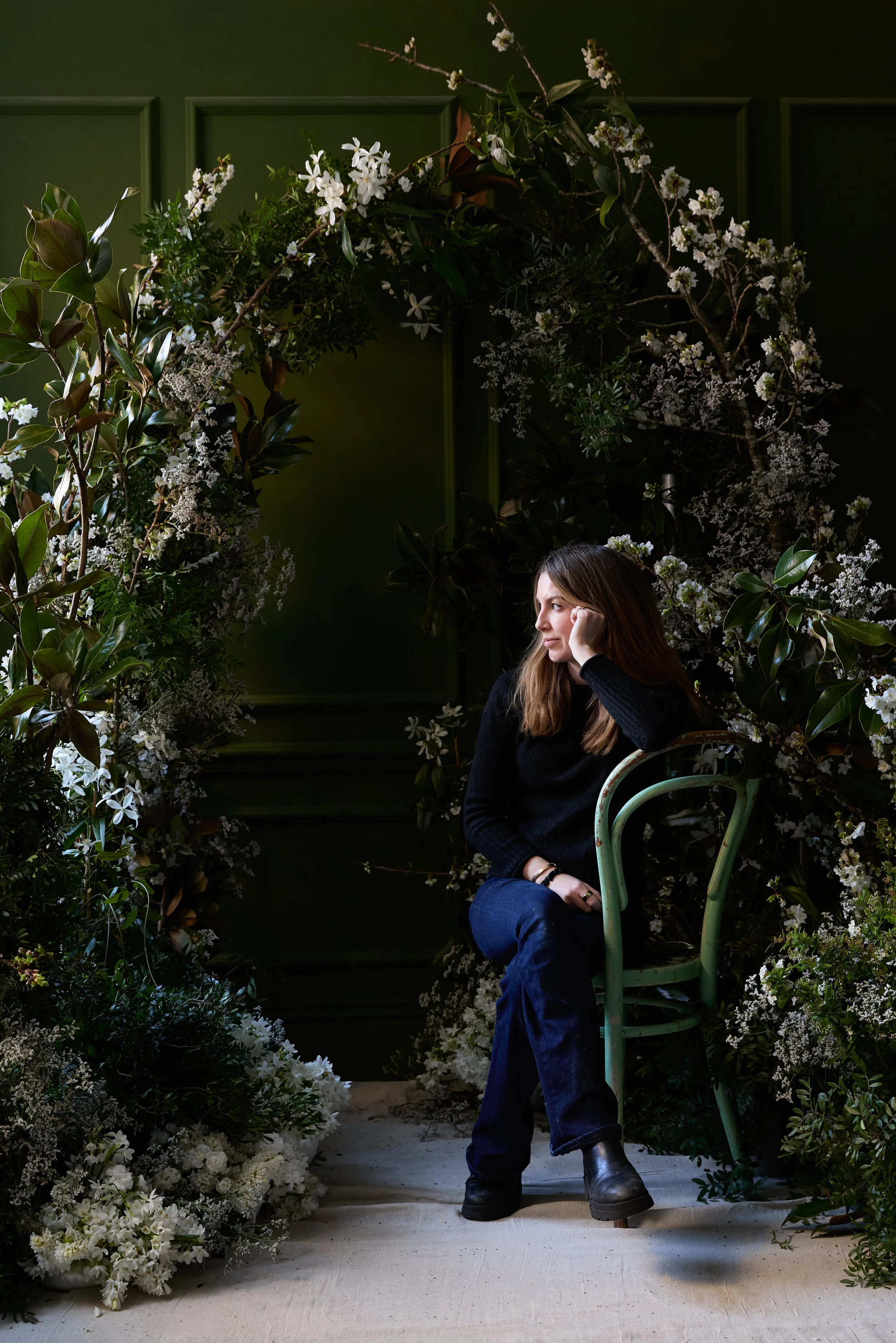A young woman with long brown hair, wearing a black sweater, blue jeans, and black boots, sits on a vintage green chair in a room decorated with lush white and green floral arrangements. She rests her head on her hand, looking thoughtfully to the side against a dark green wall.