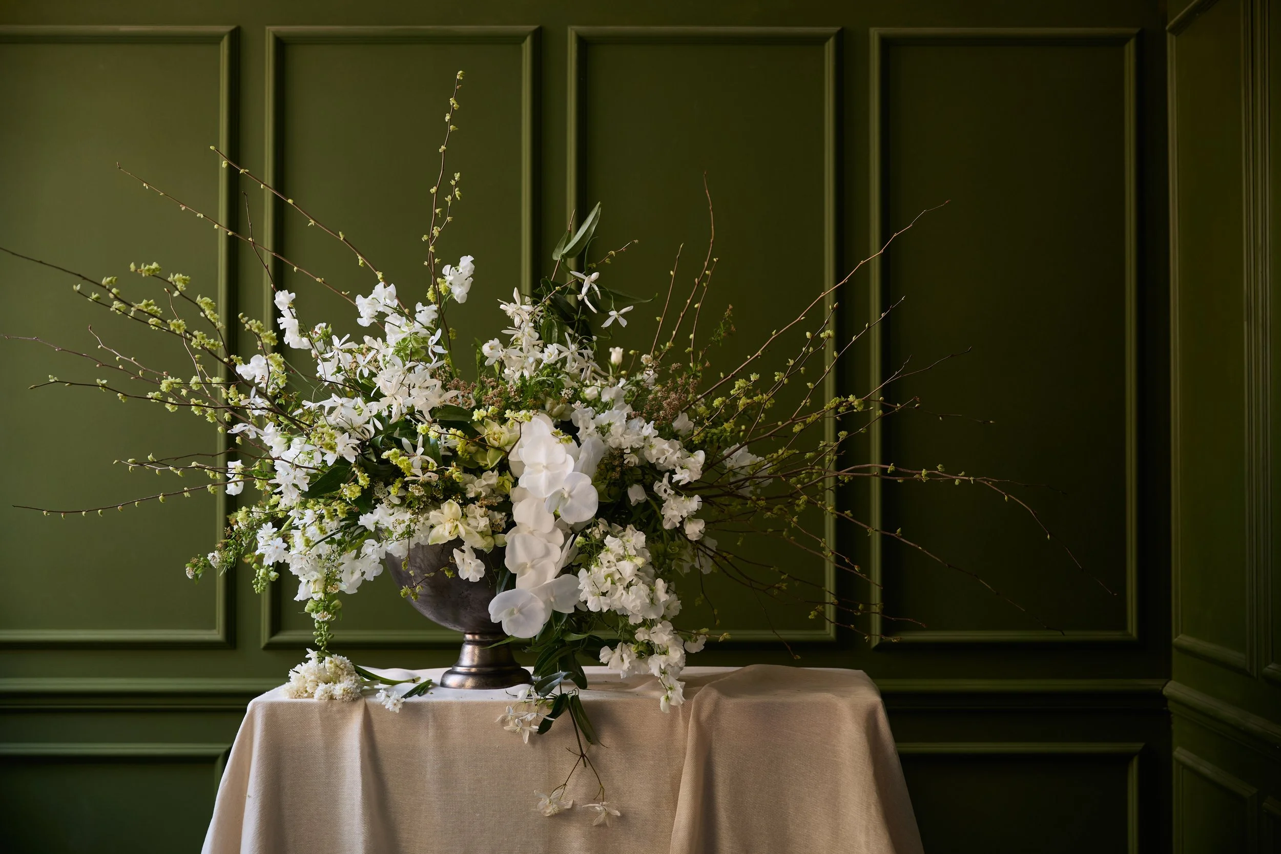 A large floral arrangement with white flowers and green foliage in a silver vase on a table with a beige tablecloth, set against a green paneled wall.