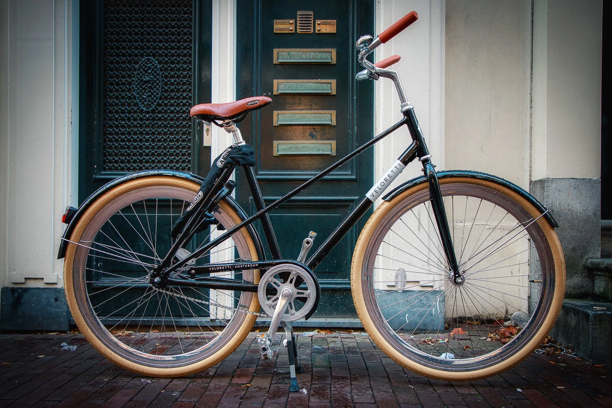 A black bicycle with tan tires leaning against a green door with mailboxes. The ground is brick and there are some leaves and debris.
