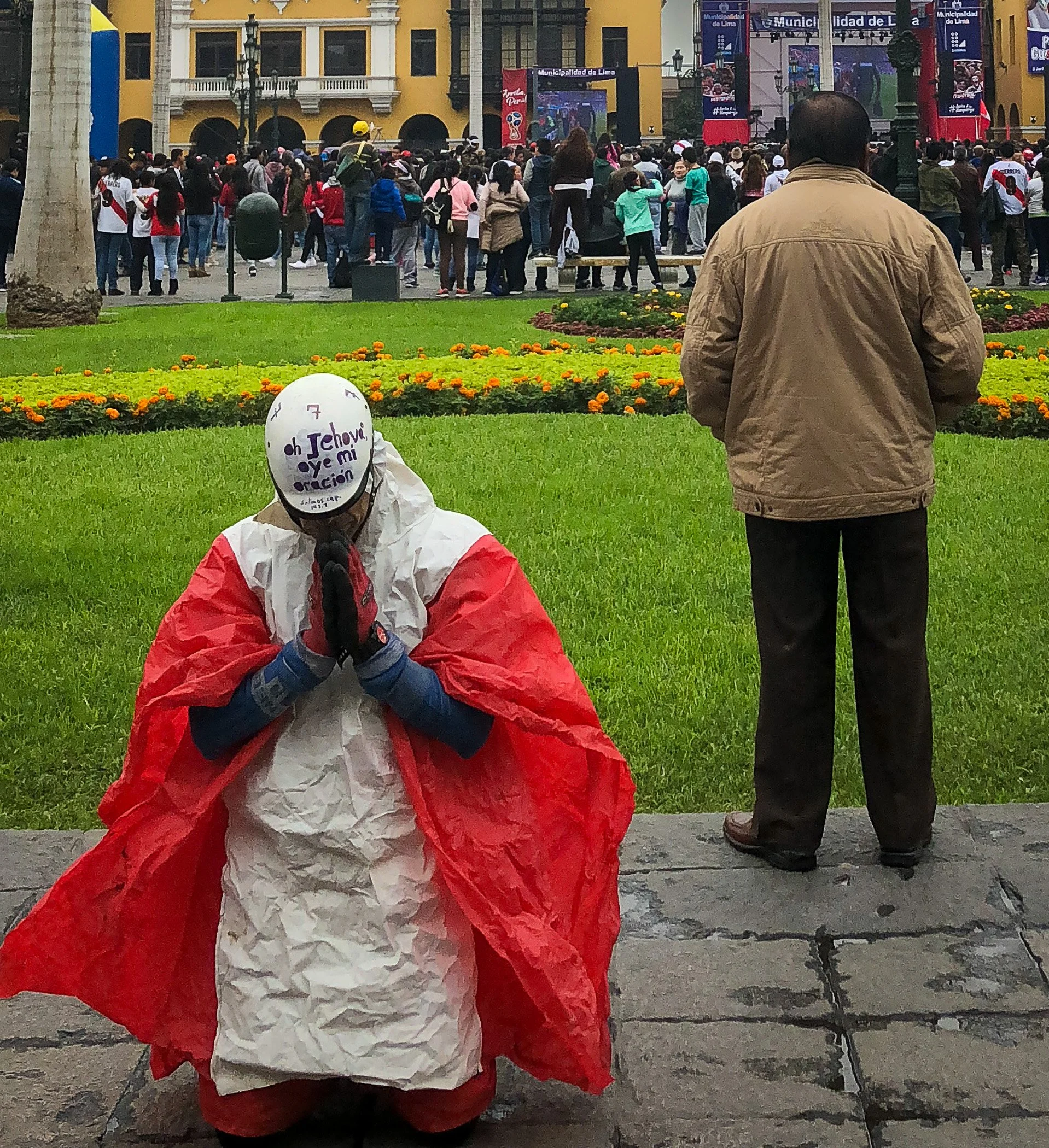 A person dressed in a costume resembling the Mexican flag with a helmet that has writing on it, kneeling with hands together in prayer, in a park with flowers and grass. Behind them, a man stands with his back to the camera, facing a large crowd gath